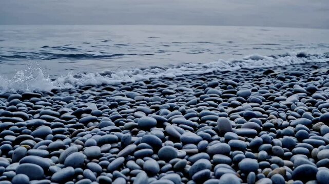 Static wide shot: scattered pebbles as waves roll in layers beyond, the cool tone color grade enhancing the tranquil mood.
