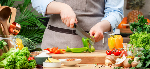 Housewife cuts green bell pepper. Cozy kitchen with wooden table, kitchenware, vegetables, herbs and ingredients for cooking. Healthy vegan eating, culinary, recipes, food blogging