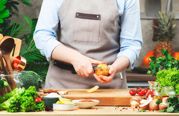 Cook peels raw potatoes with vegetable peeler. Cozy kitchen with wooden table, kitchenware, vegetables, herbs and ingredients for cooking. Healthy vegan eating, culinary, recipes, food blogging