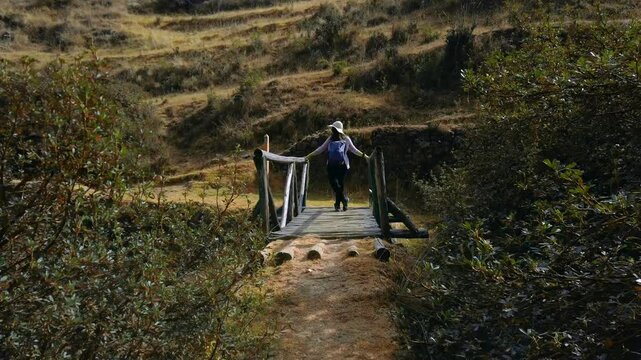 Approaching shot of a woman on a wooden bridge at Inkilltambo ruins entry. White caucasian tourist, brunette with Peruvian hat and backpack.