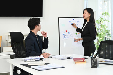 A woman is giving a presentation to a man in a suit. The woman is standing in front of a white board with a presentation on it. The man is sitting in a chair behind her.
