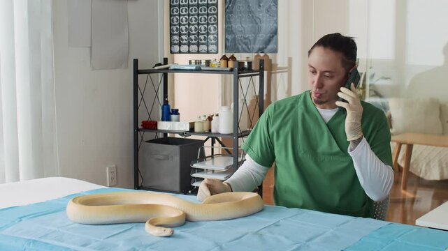 Static shot of multi-ethnic male specialist in medical uniform giving consultation on mobile phone while inspecting yellow snake on table in clinic