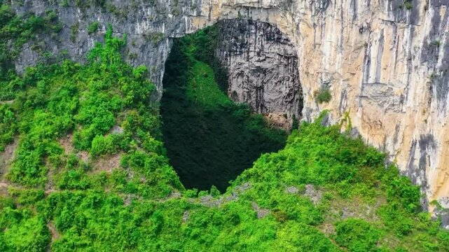 Aerial drone flying out and further into the air from Heavenly Pit of Xiaozhai Tiankeng natural sinkhole in a steep mountain cliff with green nature surrounding it in China. Sun is shining, cinematic.