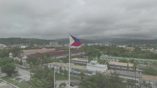 A breathtaking drone shot captures the Philippine flag waving proudly atop a vibrant green landscape, with dramatic clouds framing the scene, highlighting the nation's rich culture and natural beauty.