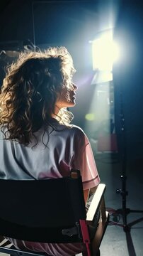 Film director sitting in a director's chair during a movie shoot, capturing the action from a back view, while the film crew works diligently in the background on the bustling cinema set