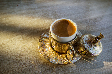 Golden Turkish Coffee Cup Set on a Stone Table. A close-up view of a traditional Turkish coffee cup and saucer set, showcasing intricate gold-toned metalwork.
