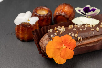 A rich chocolate ?clair with a glossy ganache layer, sprinkled with chopped nuts, and decorated with bright edible flowers, served on a dark slate surface.