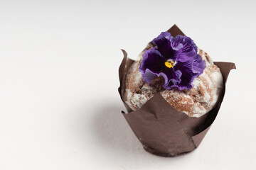 Side view of a muffin dusted with powdered sugar, wrapped in brown parchment, and adorned with a deep purple edible flower.