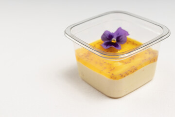 A beautifully layered mango and cream dessert in a plastic container, decorated with a striking purple edible flower.