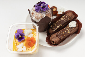 A gourmet dessert spread featuring a custard dessert, chocolate ?clairs, and a muffin, all decorated with edible flowers, presented on a white table.