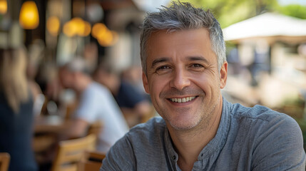 Fototapeta premium A handsome man in his late thirties is smiling at the camera. He has short gray hair and is wearing an open gray shirt, sitting outside a cafe on a summer day, generative AI
