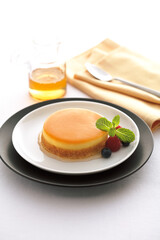 A golden cream caramel dessert put on a white plate, garnished with vibrant berries and a sprig of mint. A glass of honey and a spoon rest on a napkin in the background.