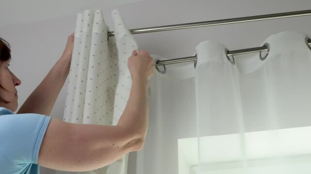 Woman hangs white curtains with metal rings made of natural material with embroidered hearts in kitchen of house. Cleaning and cleanliness. Interior and textiles. 