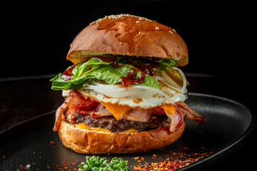 A delicious gourmet burger stacked with crispy bacon, fresh lettuce, onions, and rich sauces. Served on a sesame seed bun, it's presented on a dark plate, highlighting its appetizing layers.