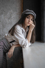Portrait of a fashionable girl in plaid trousers and a cap by the windowsill
