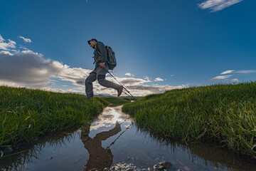 Obraz premium A tourist traveler with a backpack on his back and trekking poles jumps over a stream with green grass. A man is reflected in the water during a jump. Photo from the side of the water jump.