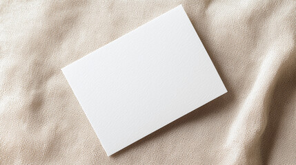 Fototapeta premium A blank white card rests on a textured beige fabric background, creating a minimalist and clean aesthetic.