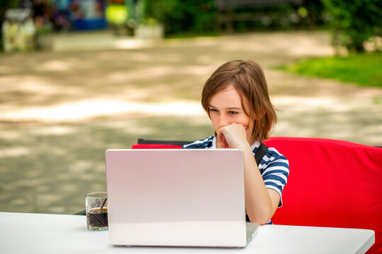 Thoughtful schoolboy studying online on computer outdoors in park. Teen boy with a laptop sits behind a table in a park. School boy is typing on computer outdoors. Child boy studying - Powered by Adobe