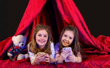 Toddler girls lies on blanket in play tent watching cartoon on tablet with interest, telephone. Little girls in play tent at night using tablet pc under blanket at night. Sisters with telephone