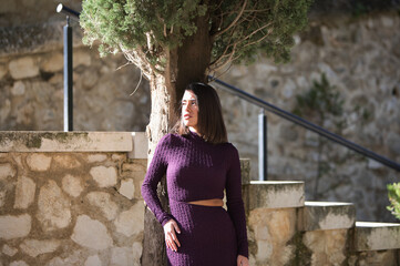 Pretty young woman dressed in elegant violet dress posing on a cypress tree by the park steps. The...