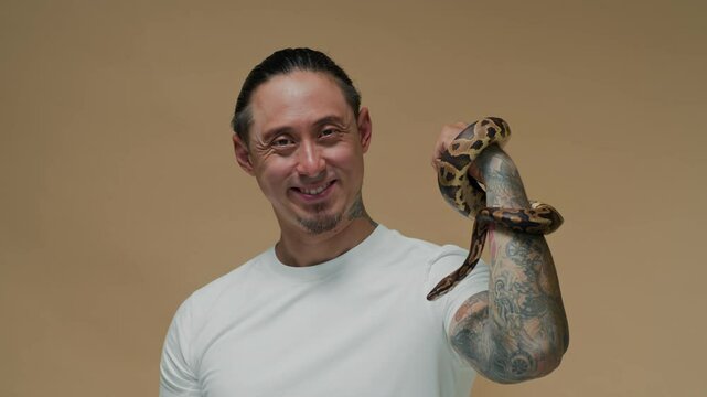 Studio portrait of cheerful multi-ethnic male owner with tattoo on arm posing for camera with small ball python, copy space