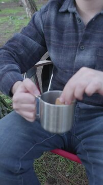 Young man with lumberjack shirt sitting on chair in forest camp and drinking coffee from metal cup