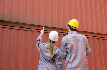 Two logistics workers in hard hats and purple uniform examining shipping container together. Woman in white helmet pointing at orange container while colleague man in yellow helmet observes at freight