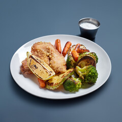 Crispy Fried Fish with Roasted Vegetables and Sauce on White Plate Against Blue Background