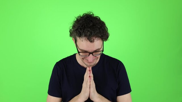 young man in dark blue T-shirt, glasses calm, happy expression greets with hands clasped in front of him. sign namaste symbol, Indian greeting. isolated on green studio background, chromakey.