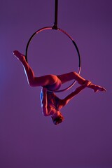 Aerial acrobat in a stunning contortion pose on a hoop, illuminated by dramatic purple lighting....