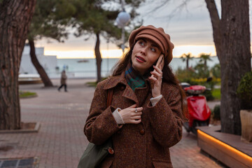 Fototapeta premium Portrait of a young beautiful Caucasian woman in a hat and coat walking down the street, smiling and talking on the phone. Concept of modern communications
