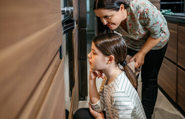 Mother and daughter are carefully watching cookies or cake baking in the oven, anticipating the delicious outcome of their work. Family time cooking concept.