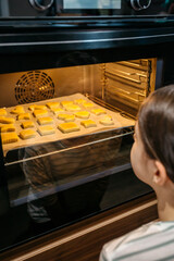 Unrecognizable little girl watching cookies baking in oven, waiting for them to be ready, enjoying the sweet smell and anticipating the delicious taste. Cooking at leisure time concept.