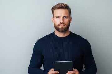 Bearded man in a navy blue sweater holding a tablet against a gray background. Modern technology and focus.