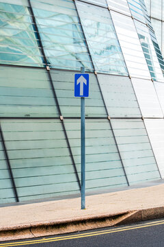 Blue directional road sign in front of a curved glass building in an urban environment.