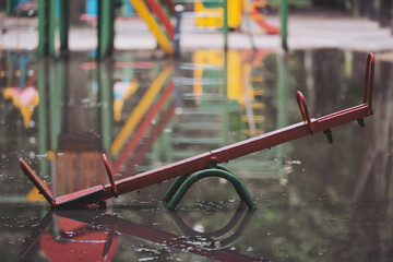 A red seesaw rests partially submerged in a large puddle, surrounded by colorful playground equipment. The gray sky mirrors in the water, creating a serene yet melancholic atmosphere