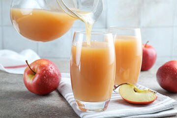 Pouring refreshing apple juice from jug into glass at grey textured table, closeup