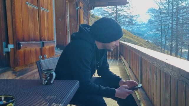 A man sits on a chalet balcony in the Swiss Alps on a crisp winter morning, smoking, playing with his phone and talking to himself
