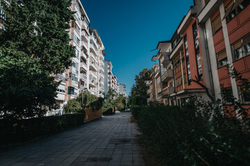 A tranquil pathway stretches between apartment buildings adorned with vibrant greenery under a clear blue sky, offering a sense of peace and urban harmony.