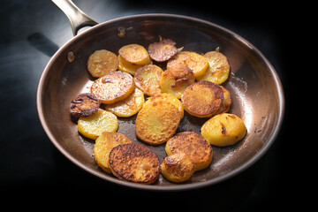 Crispy brown fried potato slices, in Germany called Bratkartoffeln, in a frying pan on a black hob in the kitchen at home, cooking concept, selected focus