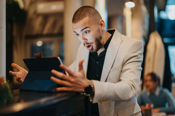 A confident businessman expressing excitement while viewing surprising news on a digital tablet in an indoor setting, symbolizing technology, communication, and business success.