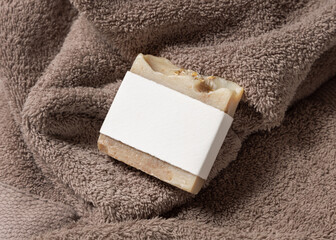 Beige herbal soap bar with blank label on light brown bath towel top view, hygiene product mockup