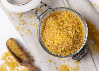 Glass jar of raw dry uncooked yellow bulgur grain with a wooden scoop on napkin top view