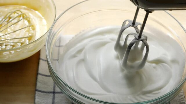 Electric Hand Mixer Whipping Cream in Glass Bowl on Wooden Countertop with Whisk and Batter in Background