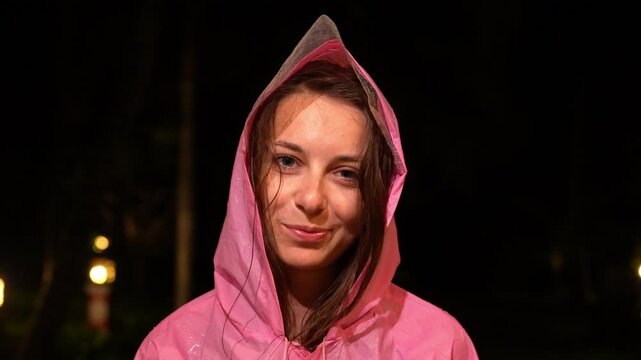 Young woman in bright pink rain jacket at night. Night portrait featuring girl in vibrant pink hooded raincoat. Pink waterproof jacket with hood highlights young woman against night backdrop. 