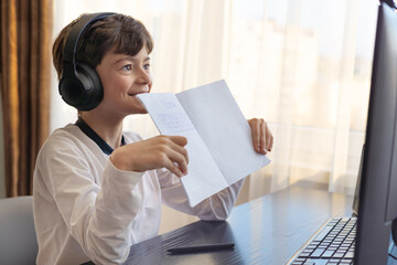 A smiling boy 7-9 years old studies online at his desk. He proudly displays his homework notebook on screen, capturing home learning, distance education, and challenges related to autism and ADHD. 