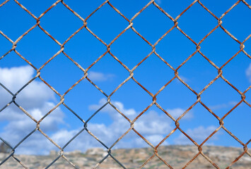 A close-up shot of a rusty wire fence against a backdrop of a clear blue sky with scattered clouds. This image symbolizes obstacles, boundaries, and the passage of time.