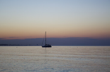 Fototapeta premium silhouette of a sailing boat, sunset by the sea, boat sunset, dramatic purple sky, gray blue clouds, lights of the coast at sunset, lonely boat, purple cloud cover, gentle waves, silhouette boat 