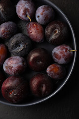 Juicy ripe fresh plum with water drops in gray plate on black background. Seasonal fruits. Selective focus, close-up.