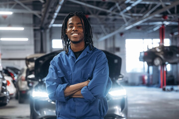 Friendly male mechanic is with car in garage, standing with arms crossed